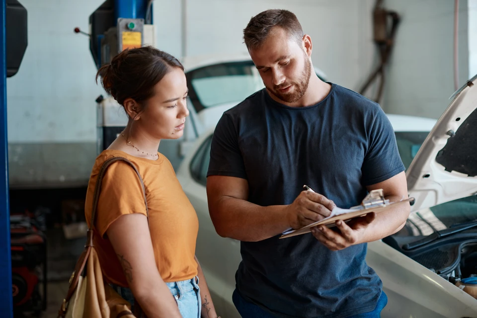 Photo professionnelle en intérieur d'atelier automobile : un véhicule utilitaire ou une berline professionnel en cours d'intervention pour réparation de vitrage, avec un technicien en tenue de travail visible effectuant une réparation ou un remplacement de pare-brise. L'environnement d'atelier moderne et bien équipé (établi, outils de réparation, système d'éclairage professionnel) doit être visible en arrière-plan pour contextualiser le cadre B2B et la prise en charge professionnelle des flottes de véhicules.